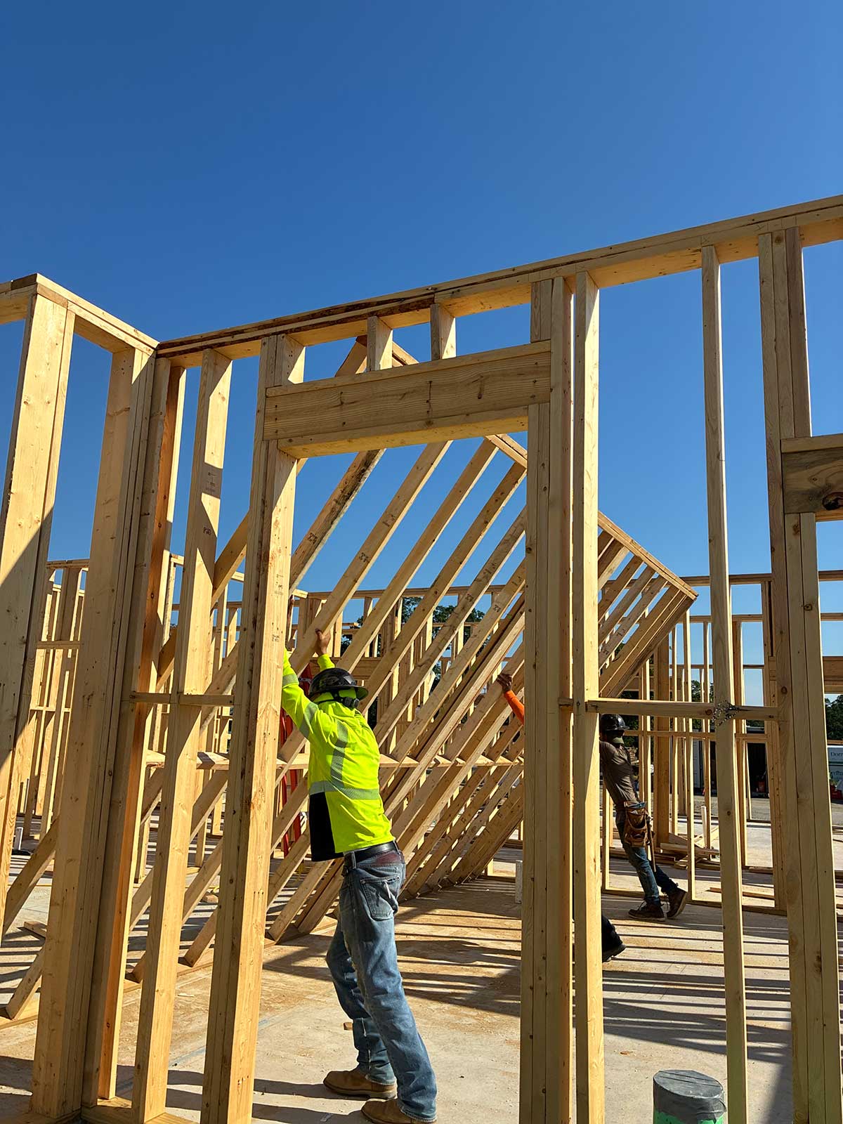 Two construction workers in safety gear raise a wooden wall frame on a building under construction, with bright blue sky in the background.