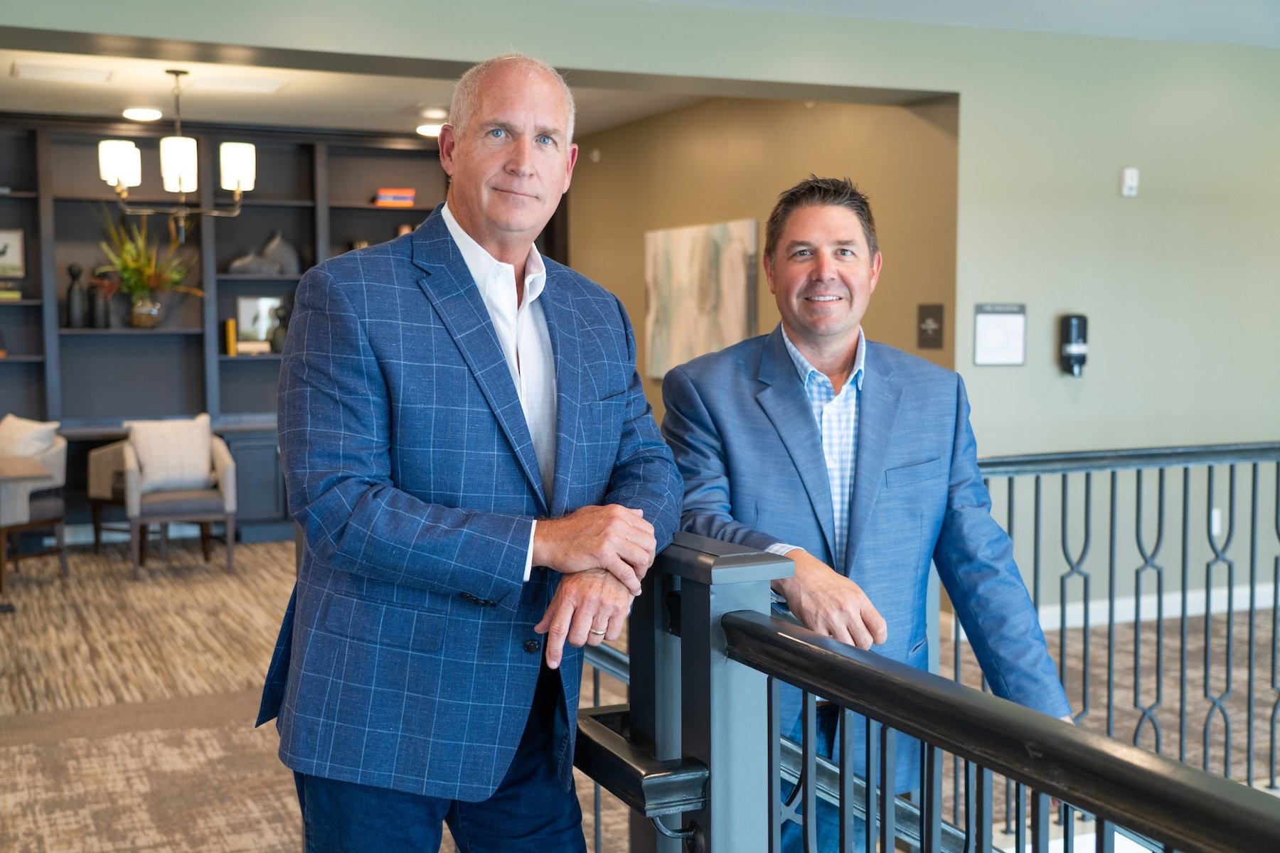 Two men in blue blazers stand by a railing in a modern office space, both looking at the camera and smiling. Shelves, chairs, and decorative items are visible in the background.
