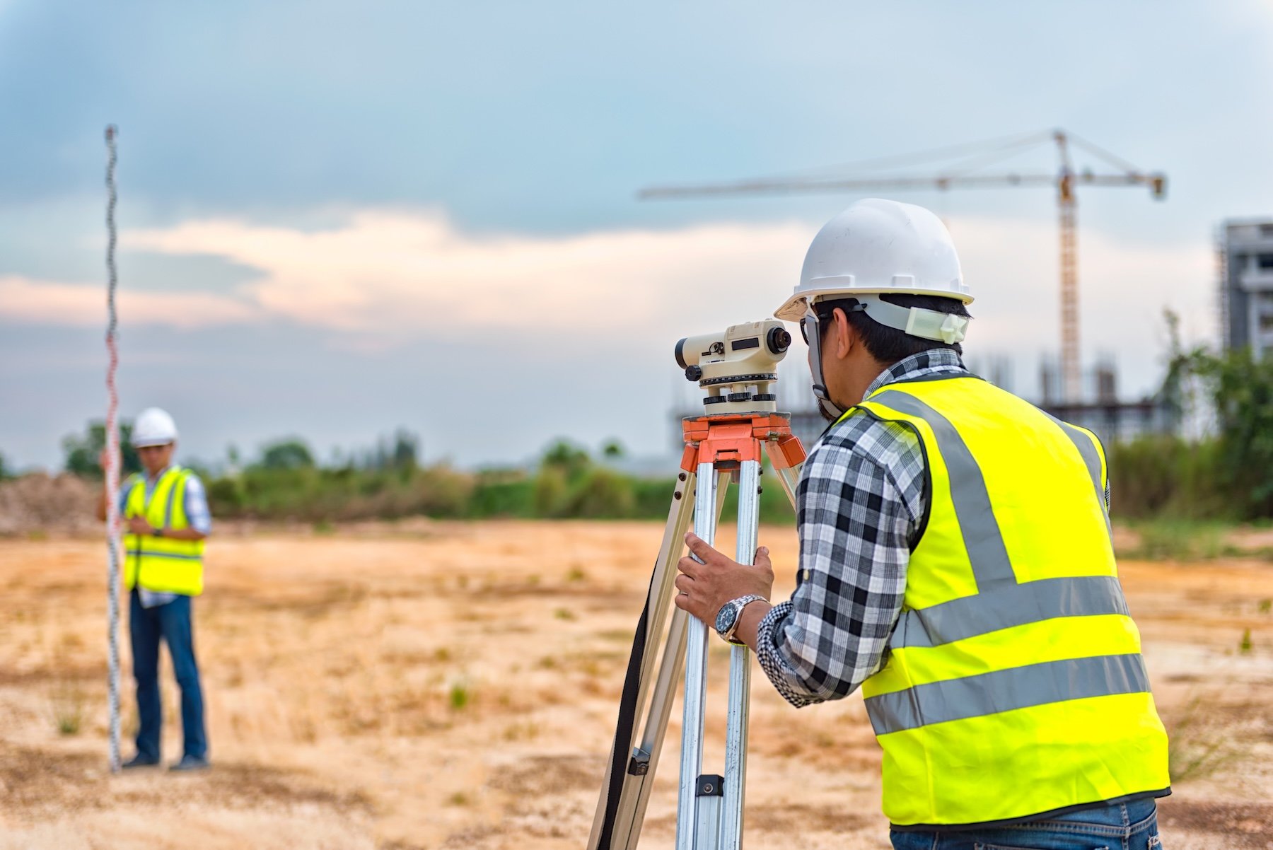 Two construction workers in safety gear raise a wooden wall frame on a building under construction, with bright blue sky in the background.